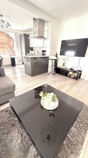 A modern living room with a black coffee table, a grey sofa, a kitchen area with a stove, and a flat-screen TV mounted on the wall. - Apartment Sleeps 11 with Air Conditioning (London)