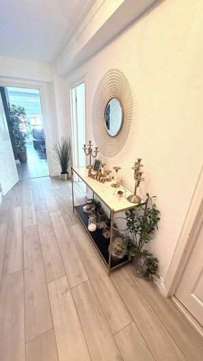 A hallway with a long table displaying various decorative items, a round mirror on the wall, and a potted plant. - Apartment Sleeps 11 with Air Conditioning (London)