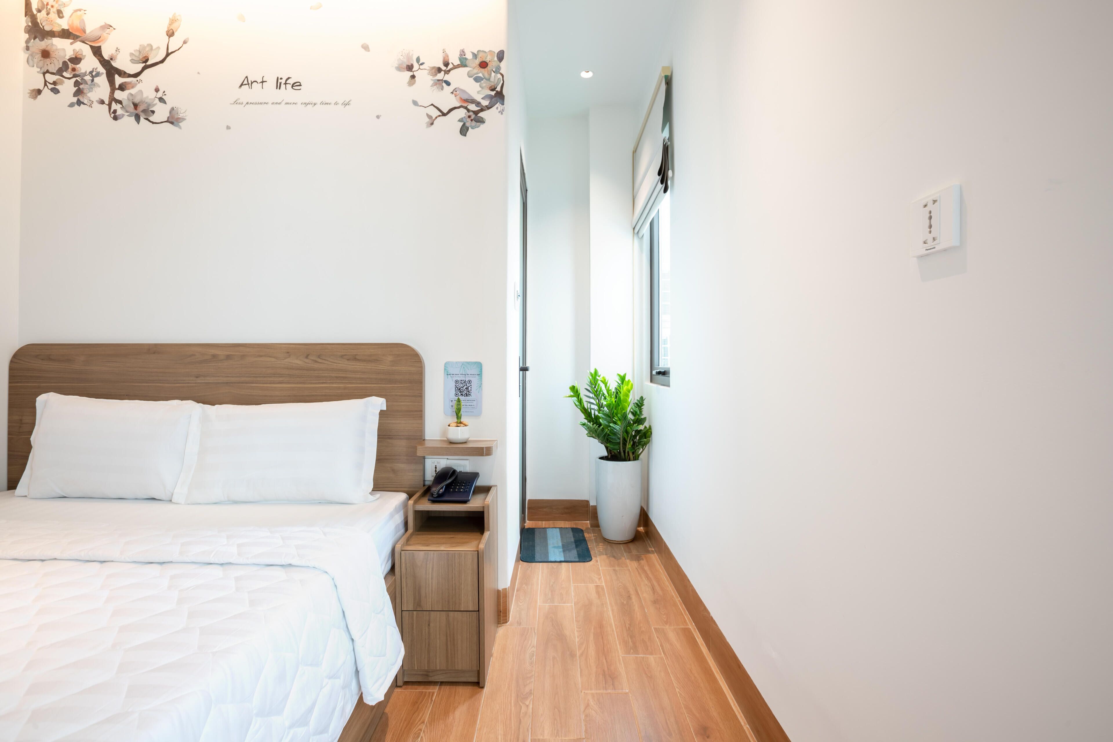 Et hotelværelse med sengegavl i træ, en seng med hvide sengetøj, et natbord med telefon og lampe, en potteplante og en væg med blomstermural og et motiverende citat.