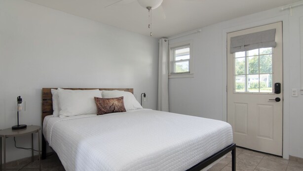A bedroom with a bed, a bedside table, a lamp, a door, and a window. - Lumber Jill Lodge Rooms (Balsam Lake)