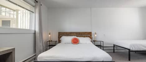 A bedroom with a large bed, two bedside tables, a wooden headboard, a window with blinds, and a ceiling light.