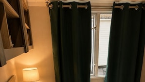 Quarto com cama, mesa de cabeceira, luminária e janela com cortinas.