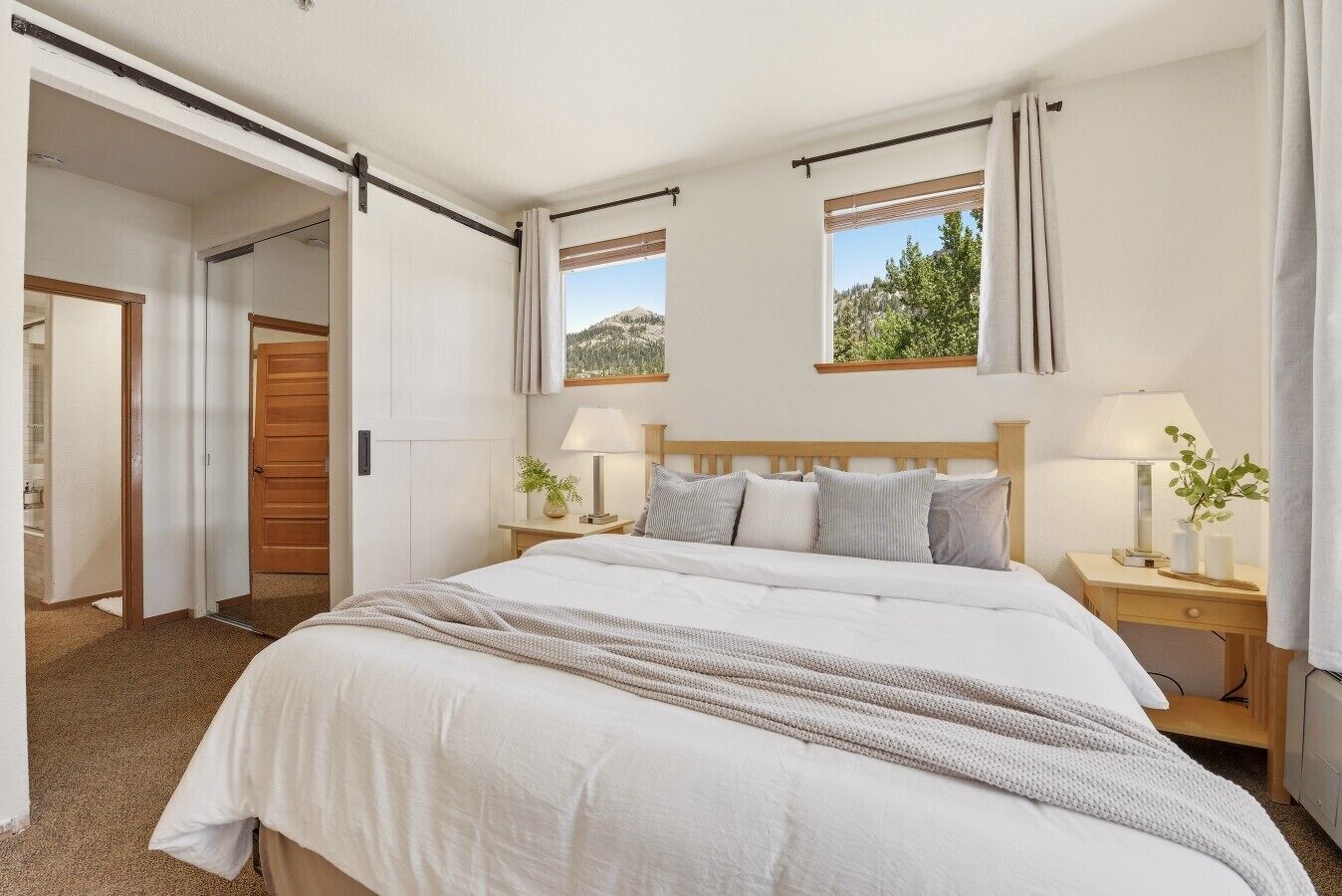Un dormitorio con una cama grande, dos mesitas de noche con lámparas, una ventana con vistas y una puerta que da a otra habitación.