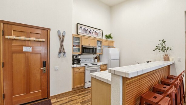A compact kitchen with wooden cabinets, a microwave, a stove, and a refrigerator. There are stools and a small table at the counter. - Mountain View Ski-In/Out Palisades Condo Hot-Tub (Olympic Valley)