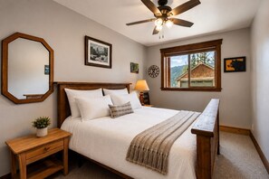 Une chambre à coucher comprenant un lit, une table de chevet, une lampe, un miroir, un ventilateur de plafond et une fenêtre donnant sur une maison.