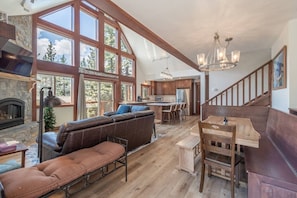 A spacious living area with a fireplace, a large window, a kitchen, and a wooden staircase.