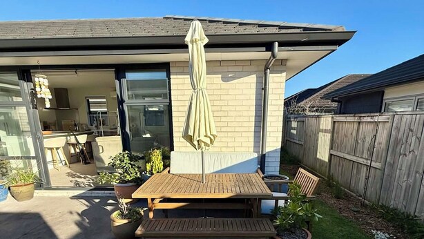 Un jardin à l’arrière d’une maison, aménagé avec une table et des chaises de patio, un parasol suspendu et des plantes en pot.