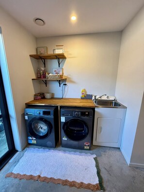 A compact laundry room with two front-loading washing machines, a sink, and wooden shelves above. - Modern 3 bedroom house in Papamoa Beach, 15 minute walk to the beach! (Tauranga)