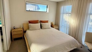 A spacious bedroom with a large bed, a nightstand, a lamp, and a view of the outside through a window.