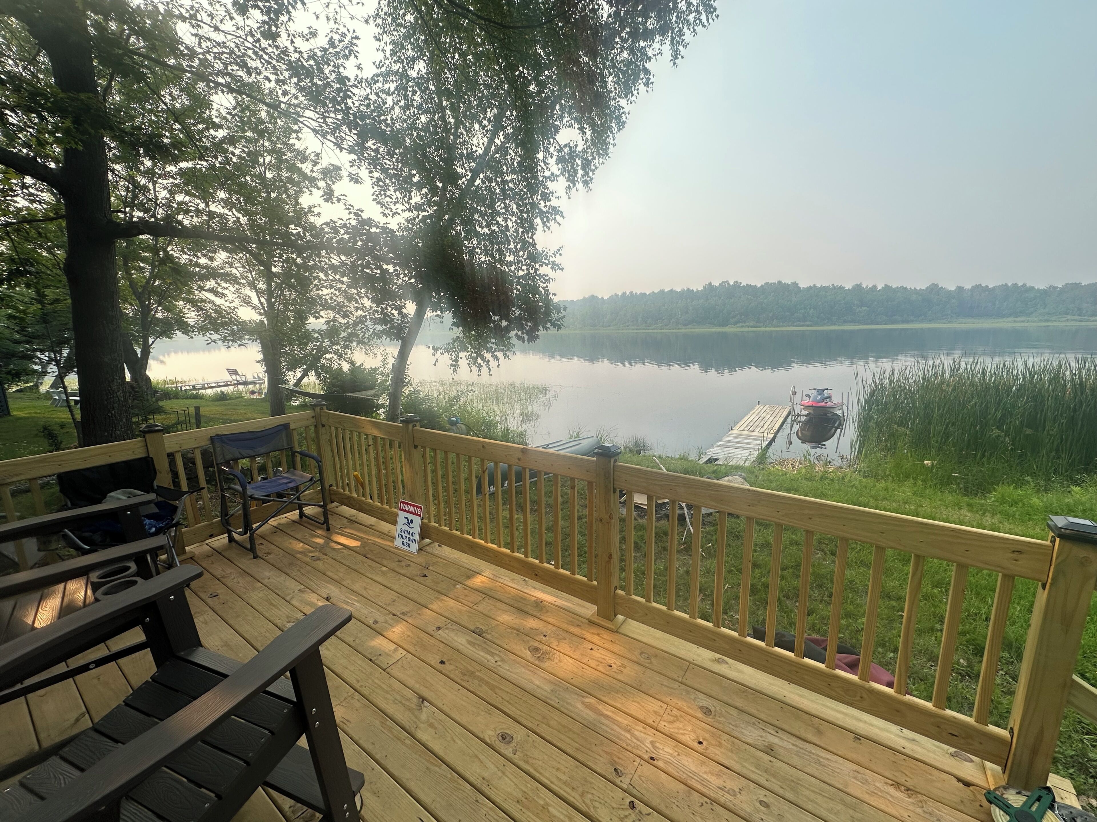 Eine Holzterrasse mit Blick auf einen stillen See, ein Steg und ein Warnschild.