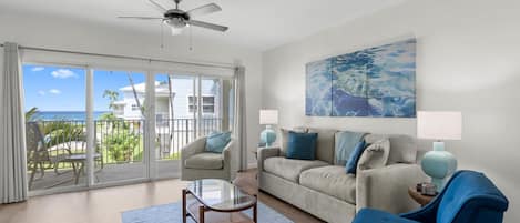 A living room with a glass coffee table, a sofa, and a chair. There is a ceiling fan, a painting on the wall, and a view of the ocean through large sliding doors.