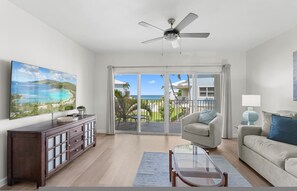 A spacious living room with a large TV, a ceiling fan, a sliding glass door to a balcony, and a sofa. - Grape Tree Villas 30 (Seven Mile Beach)