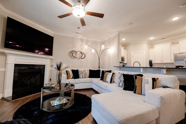 A modern living room with a sectional sofa, a fireplace, a ceiling fan, and a kitchen in the background.