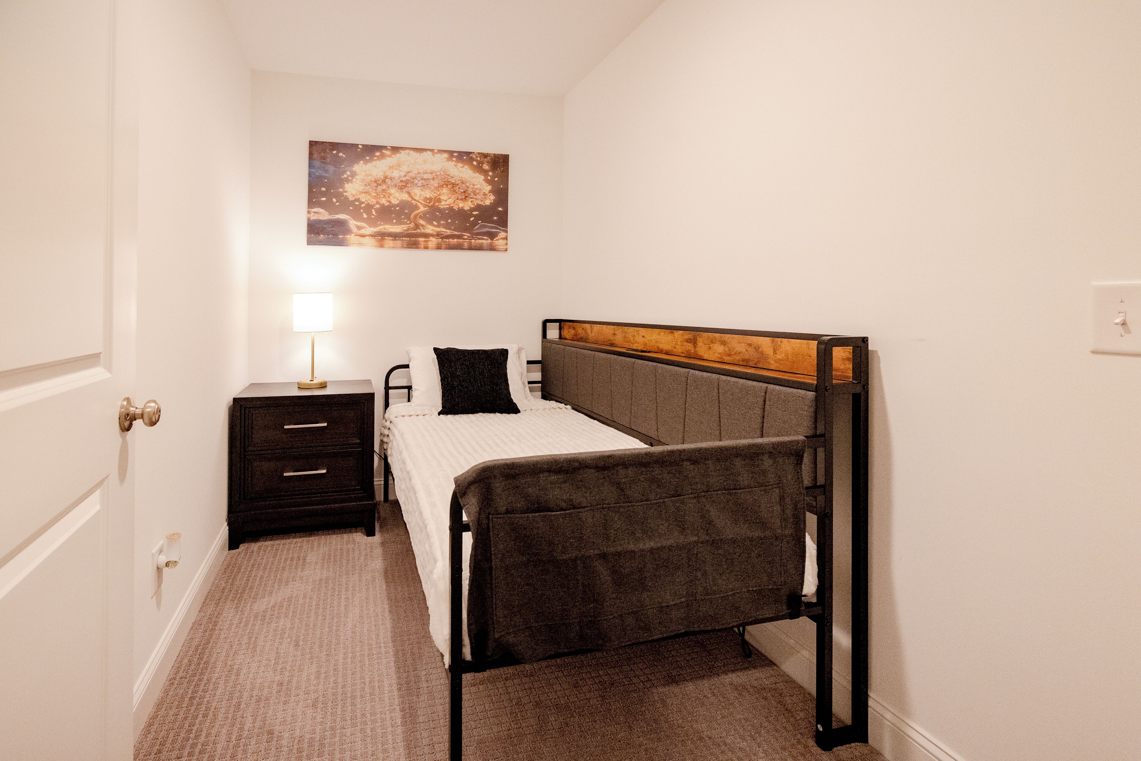 A bedroom with a single bed, a bedside table, a lamp, and a wall-mounted painting.