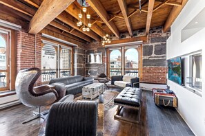 A modern living room with exposed wooden beams, a brick wall, and large windows offering city views. - Spacious & Charming 3BR - Heart of Old Montreal (Montréal)