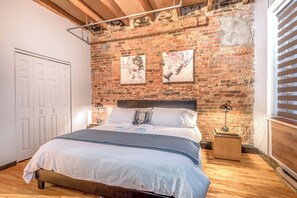 A bedroom with a brick wall, a bed with white linens, two bedside lamps, and a window with blinds. - Spacious & Charming 3BR - Heart of Old Montreal (Montréal)