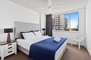 Een hotelkamer met een bed, twee nachtkastjes, een plafondventilator, een raam met uitzicht op gebouwen en een witte stoel.