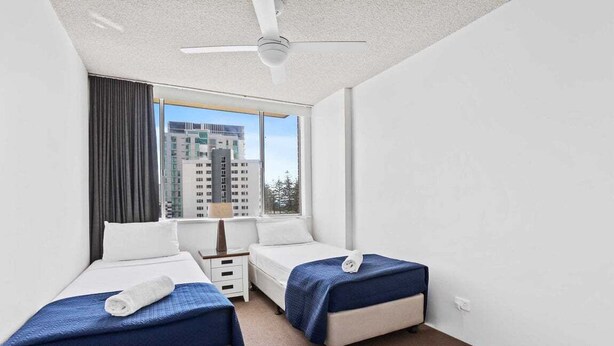 A hotel room with two beds, a ceiling fan, a window with a view of buildings, and a nightstand with a lamp. - Wyuna Ocean Views in Burleigh (Gold Coast)