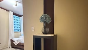 A hallway with a vase on a pedestal, a bed with a white cover, and a window with curtains.