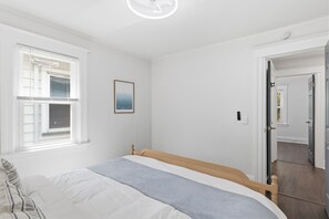 Ein Schlafzimmer mit einem Bett, einem Fenster mit Jalousien, einem gerahmten Bild an der Wand und einer Tür zu einem anderen Raum.