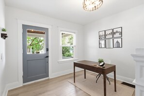 A room with a gray door, a window, a table with a clock, and framed pictures on the wall.
