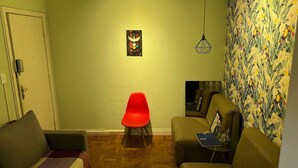 A living room with a red chair, a sofa, and a wall with a tropical print. - Quarto Individual Augusta Paulista Frei (São Paulo)