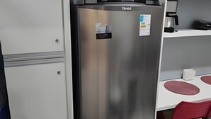 Uma geladeira de aço inoxidável em uma cozinha com um armário branco, uma cafeteira e uma cadeira vermelha.