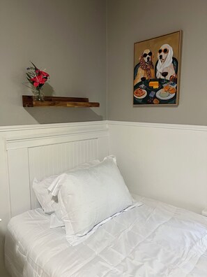 Ett sovrum med en säng, en vit kudde, en trähylla med en vas med blommor och en tavla på väggen.
