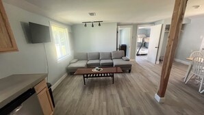 A living room with a sofa, a coffee table, and a television. There is a kitchen area with a dishwasher and a dining table with chairs. - Adorable 2-bedroom house with AC in charming Lusby (Lusby)