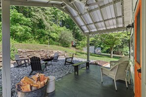 Ein überdachter Außenbereich mit Adirondack-Stühlen, einer Feuerstelle und Blick auf einen bewaldeten Hügel.