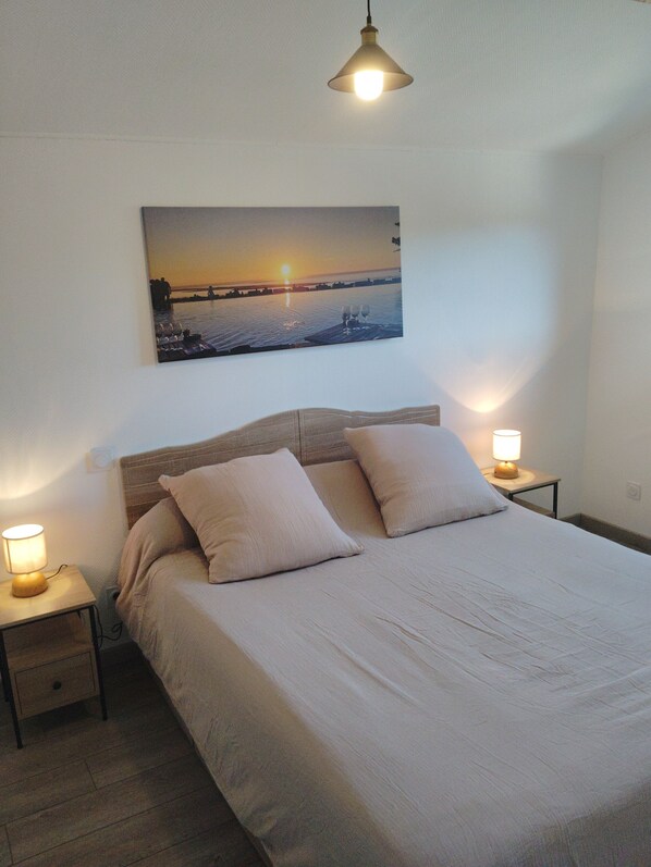 A bedroom with a bed, two bedside lamps, a nightstand, a wall-mounted picture of a sunset, and a hanging light fixture.