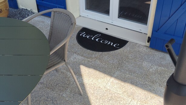 A patio area with a table and chairs, a welcome mat, and a sliding glass door leading to a vehicle. - The garleche (Sainte-Eulalie-en-Born)