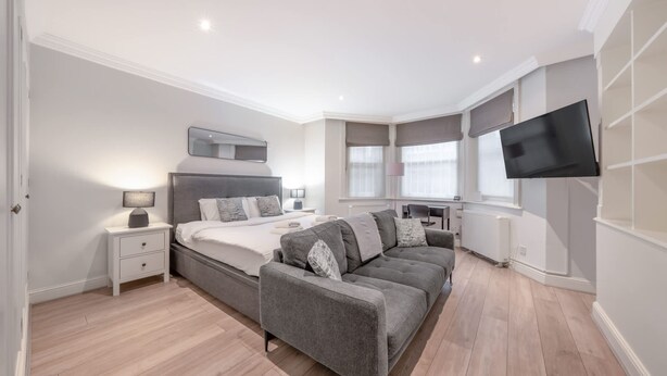 A modern bedroom with a bed, bedside tables, a sofa, a TV, and a window. - Comfort Studio by Harrods (London)