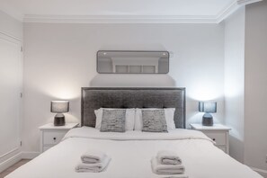 A modern bedroom with a large bed, two bedside tables, a headboard, and a mirror above the bed. - Comfort Studio by Harrods (London)
