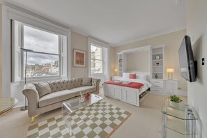 Ein modernes Schlafzimmer mit einer Couch, einem Bett, einem Glastisch und Blick auf die Stadt durch große Fenster.