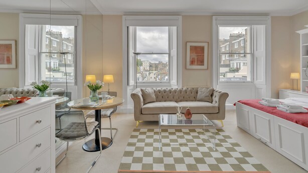 A modern living room with a sofa, coffee table, and dining area, large windows with city views, and a checkered rug. - Bright Studio in Belgravia With Views (London)