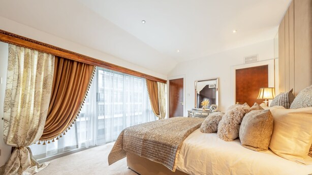 A spacious bedroom with a large bed, a bedside table with a lamp, and a view of the city through large windows. - Spacious Penthouse Near Hyde Park London (London)