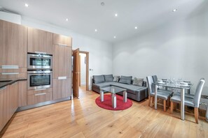 A modern kitchen with built-in ovens and a dining area with a glass table and grey upholstered chairs. - Contemporary 1B Apartment - Paddington St (London)