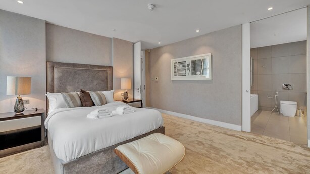 A modern bedroom with a large bed, bedside tables, a nightstand, a wall-mounted artwork, and a bathroom visible through an open door. - Beautiful Apartment With Lift in Baker St (London)