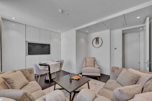 Ein modernes Wohnzimmer mit einer Sofagarnitur, einem Couchtisch, einem Esstisch mit Stühlen, einem an der Wand montierten Fernseher und einer Tür.