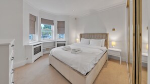 A bedroom with a large bed, bedside tables with lamps, a window with blinds, and a radiator. - Charming 2-bedroom Apartment in Knightsbridge (London)