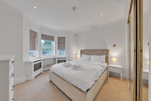 A bedroom with a large bed, bedside tables with lamps, a window with blinds, and a radiator.