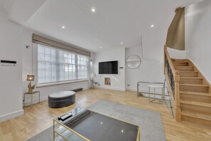 A spacious living room with a large window, a flat-screen TV, a round mirror, a staircase, and a modern coffee table. - Charming Mayfair Home With Modern Comforts (London)