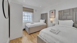 A modern bedroom with two beds, a large window with blinds, a bedside table with a lamp, and a framed picture on the wall. - Charming Mayfair Home With Modern Comforts (London)