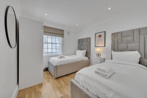 A modern bedroom with two beds, a large window with blinds, a bedside table with a lamp, and a framed picture on the wall. - Charming Mayfair Home With Modern Comforts (London)