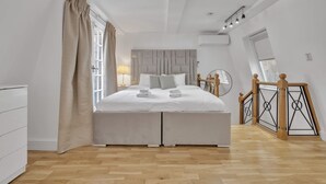 A modern bedroom with a large bed, a bedside table, a lamp, a window with curtains, and a wooden staircase. - Charming Mayfair Home With Modern Comforts (London)