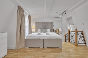 A modern bedroom with a large bed, a bedside table, a lamp, a window with curtains, and a wooden staircase. - Charming Mayfair Home With Modern Comforts (London)