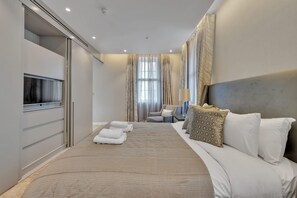 Ein Hotelzimmer mit einem großen Bett, einem Fernseher, einer Sitzecke mit Sessel und Tisch sowie einem Fenster mit Vorhängen.