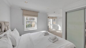 Ein Hotelzimmer mit einem großen Bett, einem Fenster mit Vorhängen, einer Tür zu einem anderen Raum und Blick ins Freie.