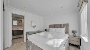 Ein modernes Schlafzimmer mit einem großen Bett, Nachttischen, einem Schminktisch und einem Spiegel. Ein Badezimmer ist durch eine geöffnete Tür sichtbar.